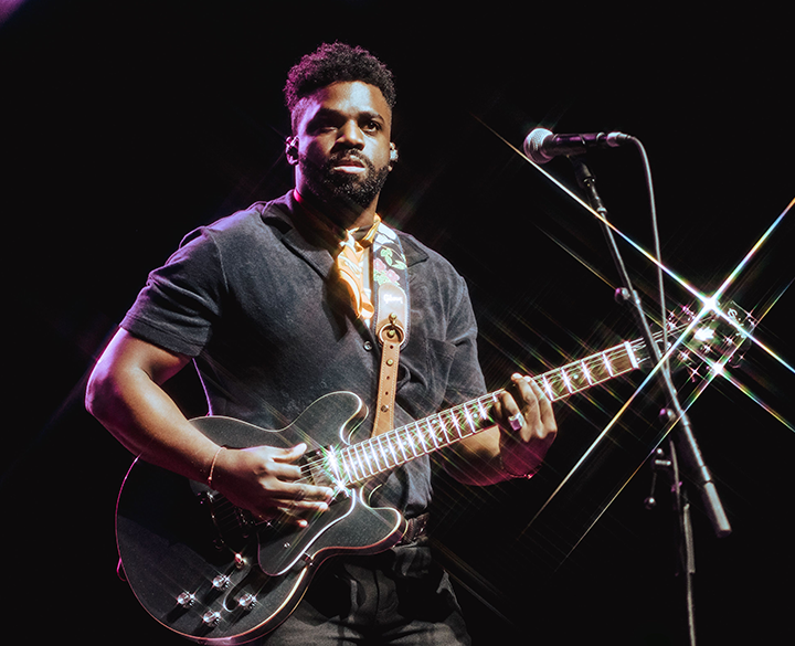 A Black man with a beard and short curly hair is playing a dark electric guitar on stage, looking intently towards the left. He is wearing a dark short-sleeved shirt, dark pants, and a patterned yellow bandana around his neck, with a guitar strap over his right shoulder. A microphone stand is visible behind him to the right, and bright stage lights create starburst effects around the headstock of his guitar.