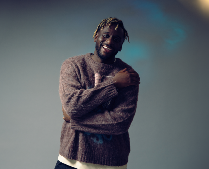 A studio portrait of a man with blonde-tipped dreadlocks wearing a fuzzy, maroon-brown patterned sweater over a light-colored undershirt. He is posing with his arms crossed over his chest, hugging himself, and smiling broadly at the camera. He is illuminated by studio lighting against a plain gray background with a hint of blue light on the right.