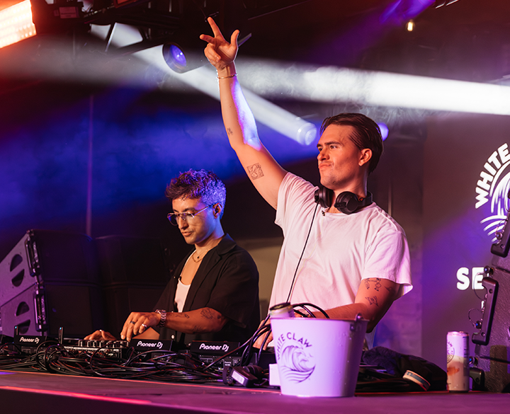 A photo of two men DJing a set in a club or on a small stage. The man on the right is energetically engaging with the crowd, raising his hand, while the man on the left is mixing music on the Pioneer decks. Bright white and purple stage lights illuminate the performers from behind.